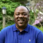 Alumnus Donté Gibbs smiles at the camera, wearing a royal blue polo shit, standing outside
