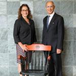 Lia Logio and Stan Gerson stand behind a CWRU-branded chair while wearing formal attire and smiling at the camera in front of a gray marble tile wall