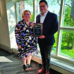 Laura McNally and Andrew Geronimo smile at camera while wearing formal attire near a window where sunlight passes through. Andrew is holding an award plaque.