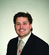George Hudak smiles at camera in front of a plain portrait backdrop while wearing a suit and tie.
