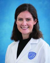 Landry Cowles smiles at camera in front of a plain portrait backdrop while wearing a medical lab coat