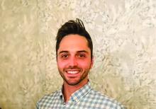 Kyle Stagg smiles at camera in front of a wall while wearing a collared shirt