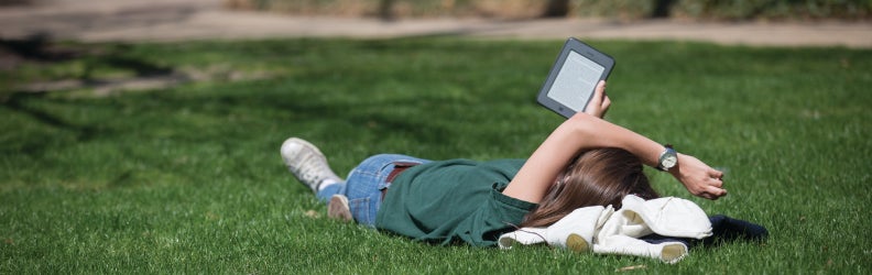 Student reading eBook lying on grass in Quad, summer time