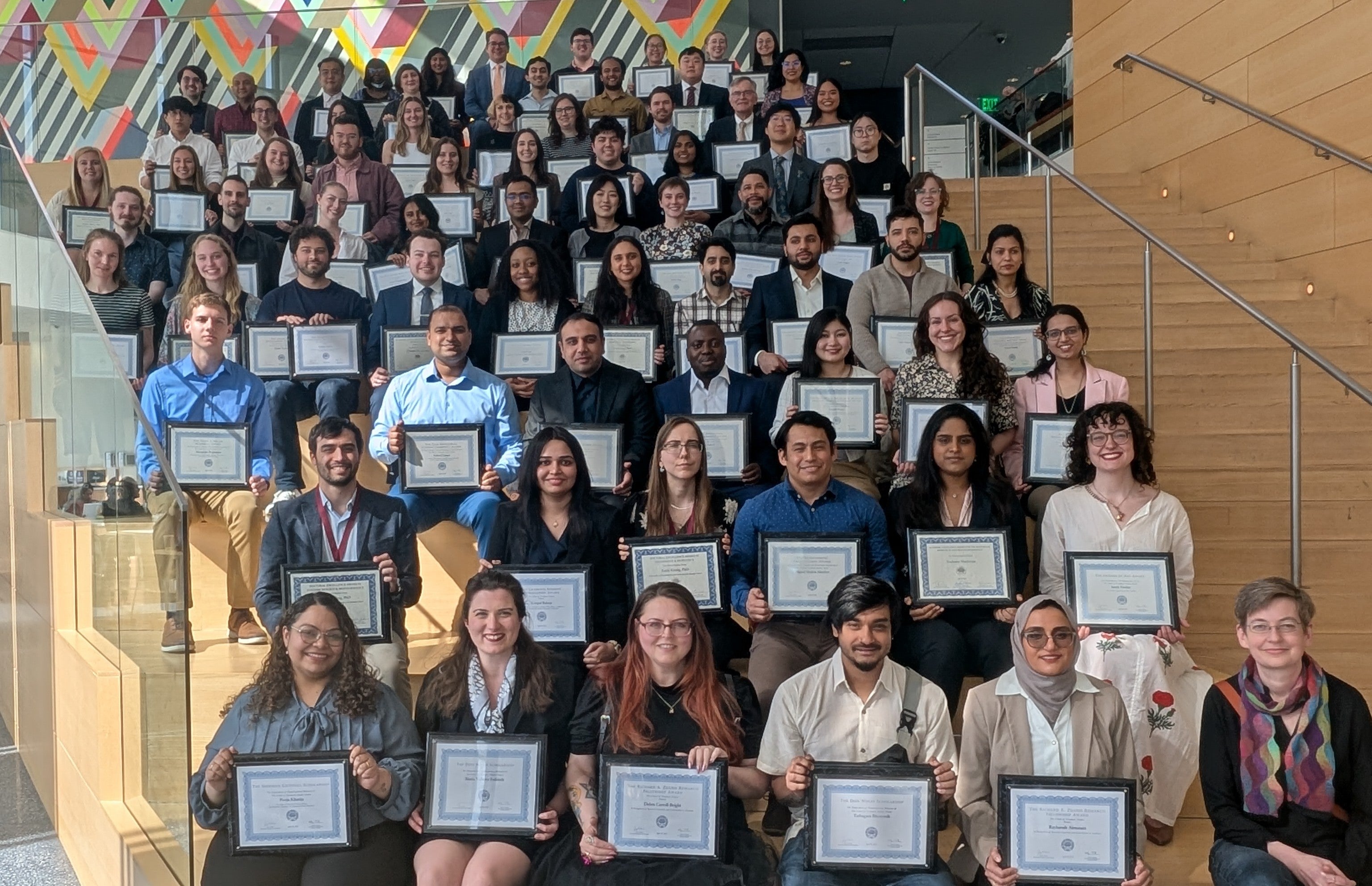 Dozens of graduate student award winners holding their plaques on the steps in the common area of Tinkham Veale University Center.