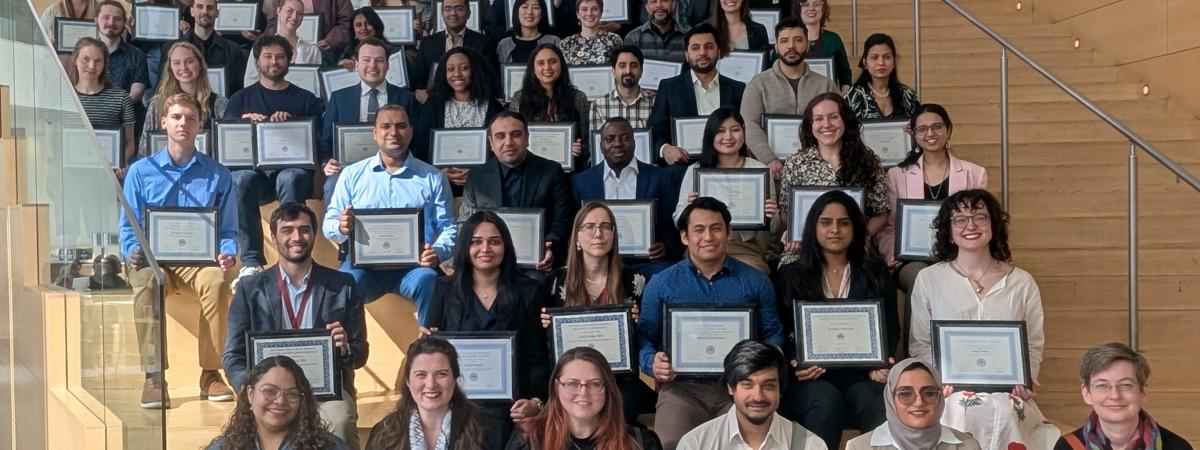 Dozens of graduate student award winners holding their plaques on the steps in the common area of Tinkham Veale University Center.