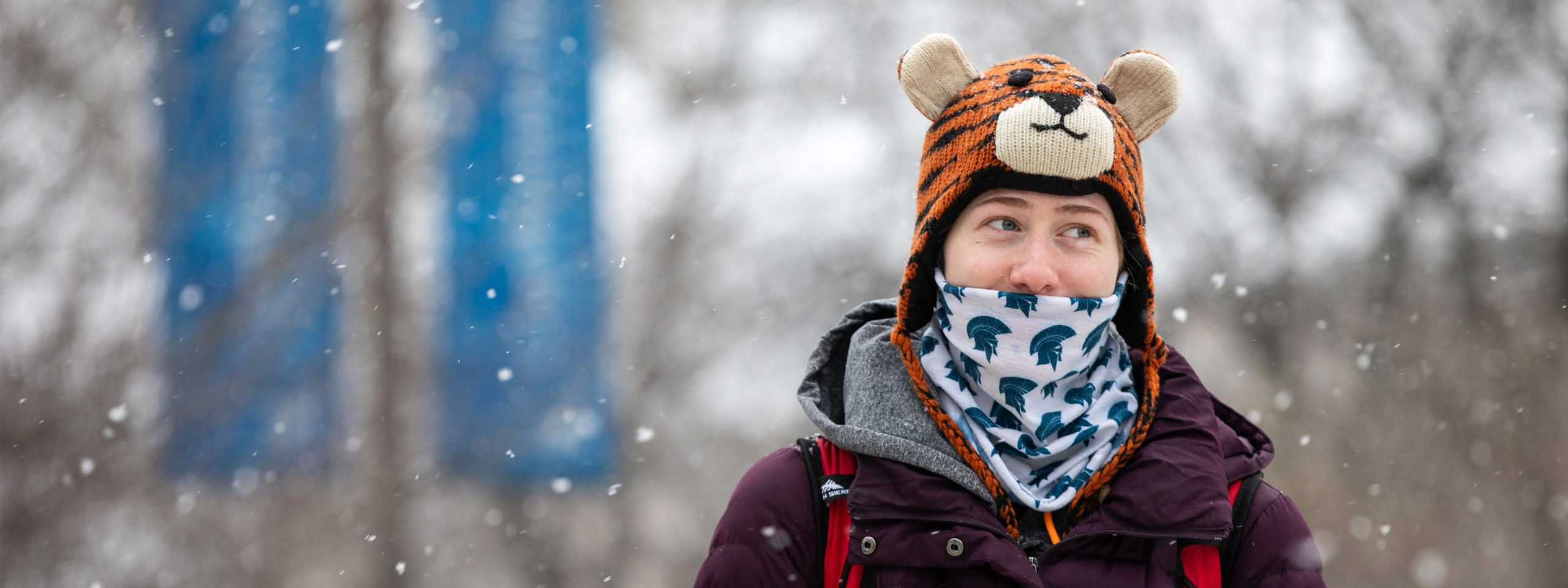 Photo of a student walking across campus on a snowy day with a bandana printed with the Spartan logo in front of her face