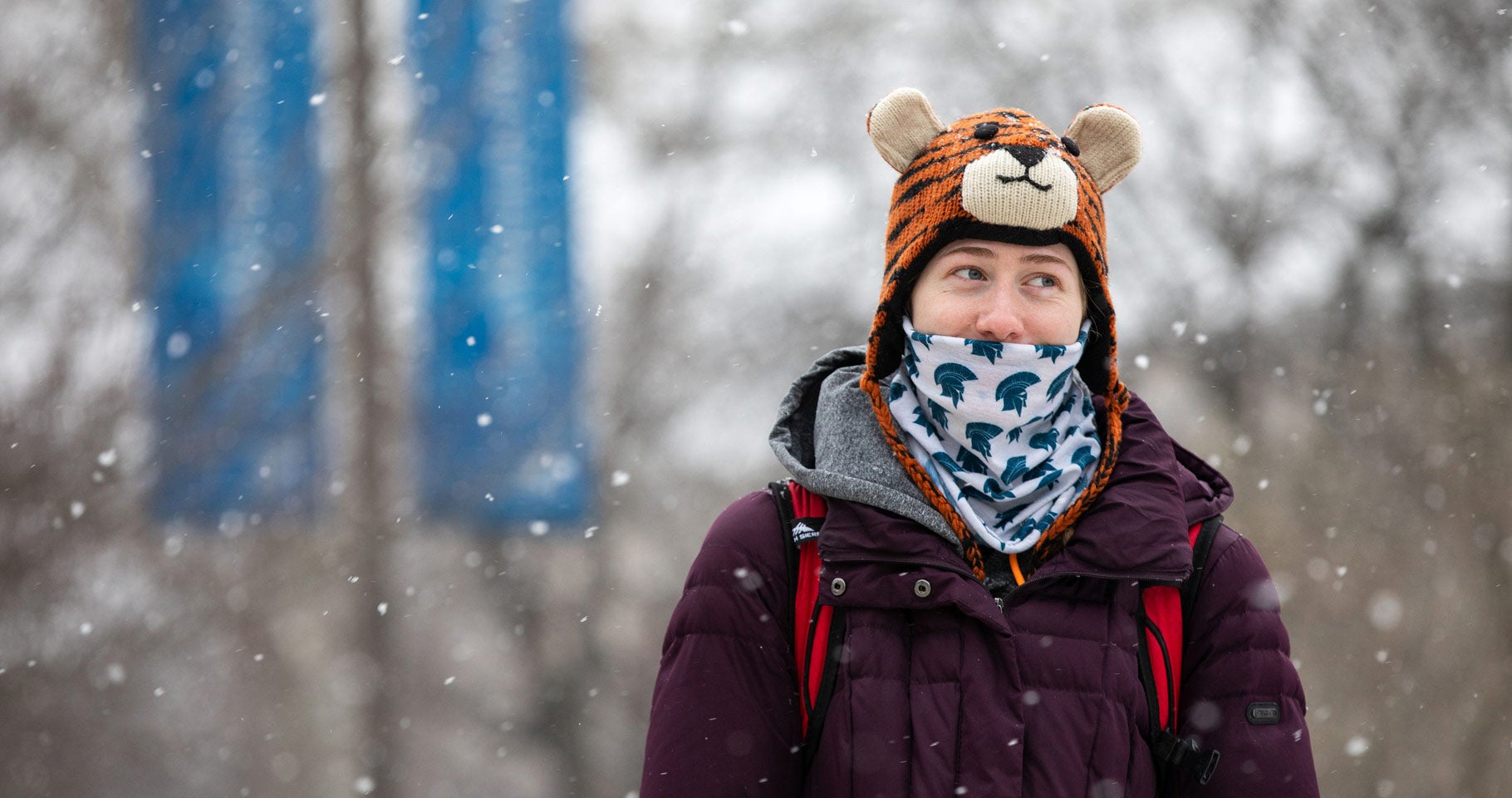 Photo of a student walking across campus on a snowy day with a bandana printed with the Spartan logo in front of her face