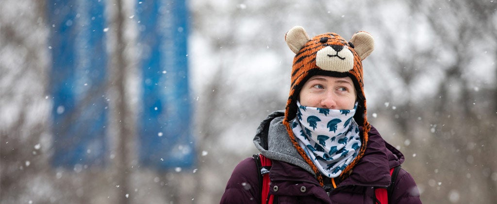 Photo of a student walking across campus on a snowy day with a bandana printed with the Spartan logo in front of her face