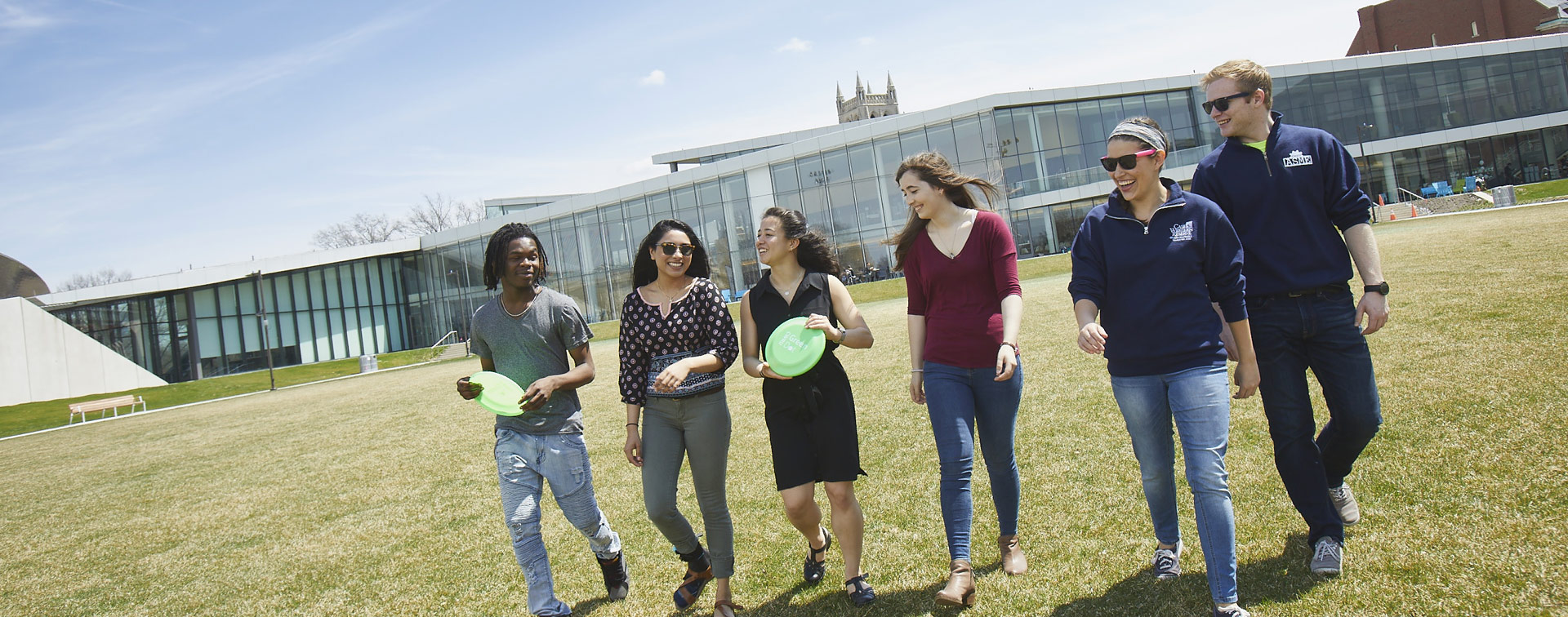 Photo of six smiling students walking across a green space with Tinkham Veale University Center behind them