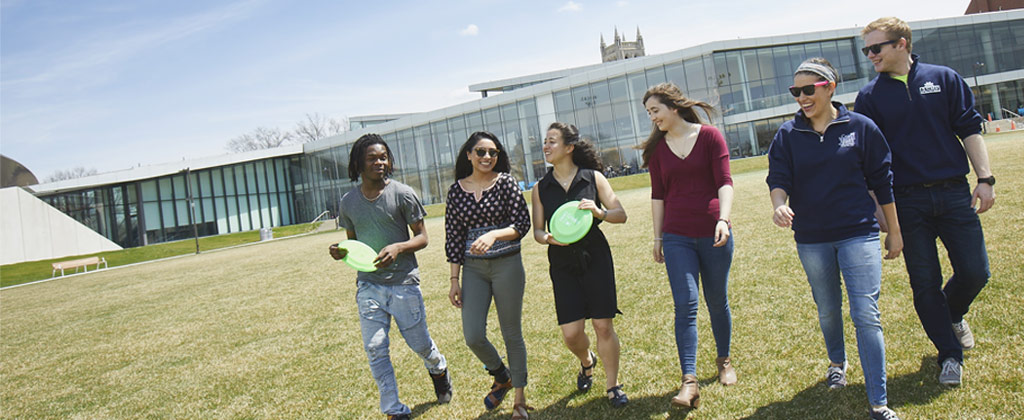 Photo of six smiling students walking across a green space with Tinkham Veale University Center behind them