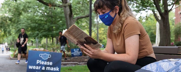 Photo of someone sitting outdoors on CWRU campus wearing a mask with a sign in the background that says please wear a mask