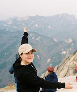 A picture of Anna Koch standing on a mountain looking at the camera with her arms extended as if to frame the mountains behind her