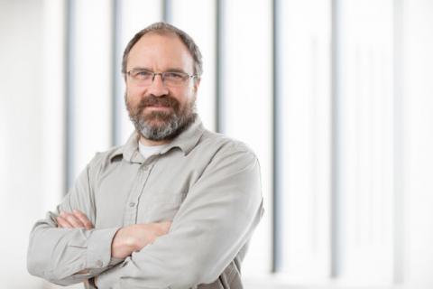 Dr. Andrew Rollins stands facing the camera with his arms crossed in front of a background of windows