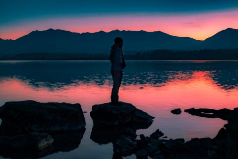 A student stands on a rock in water with mountains behind it at sunrise