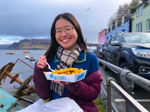 A student sits on a waterfront in Scotland holding fish and chips 