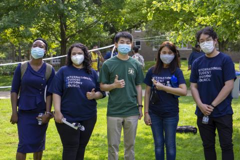 A group of international students stand together outside for a picture, some wearing shirts that read "International Student Advisory Council"