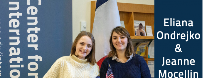 A picture of Eliana Ondrejko and Jeanne Mocellin standing in front of a French flag, each holding a flag from their home country. Jeanne is holding the French flag and Eliana is holding the American flag.