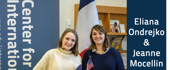 A picture of Eliana Ondrejko and Jeanne Mocellin standing in front of a French flag, each holding a flag from their home country. Jeanne is holding the French flag and Eliana is holding the American flag.