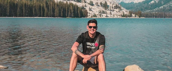 A picture of Márcio O’Dwyer sitting outside on a rock in front of a body of water with trees and mountains in the background