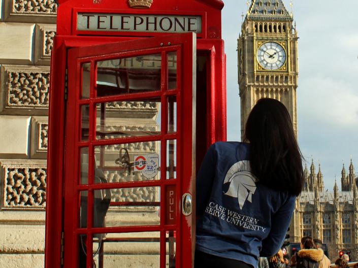 A female student in a CWRU shirt peeks out of a red telephone booth in London with Big Ben in the background