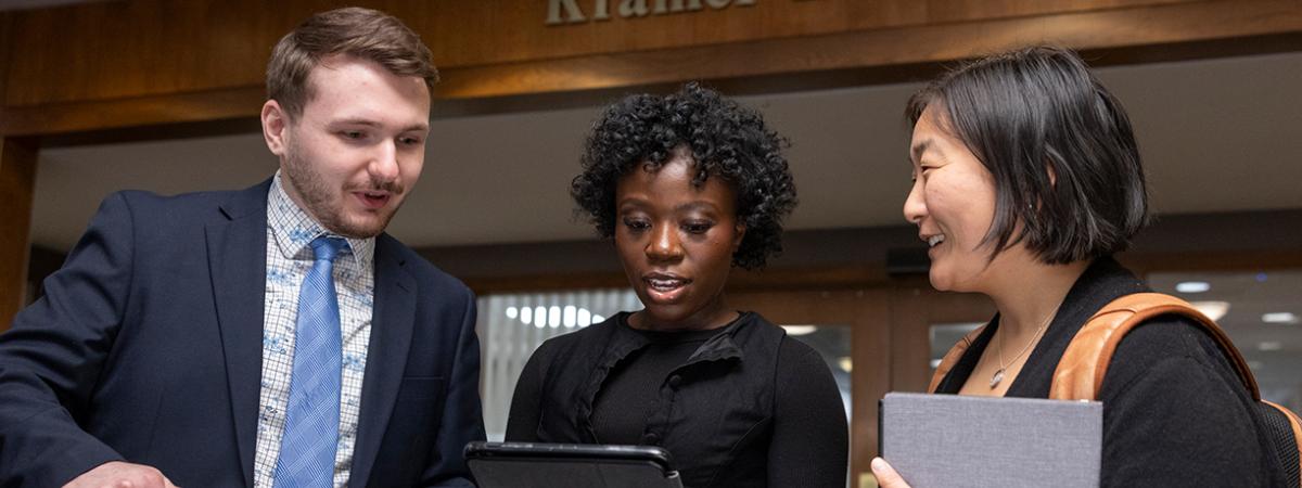 Three law students in professional attire collaborating on a project using digital devices in a campus building.