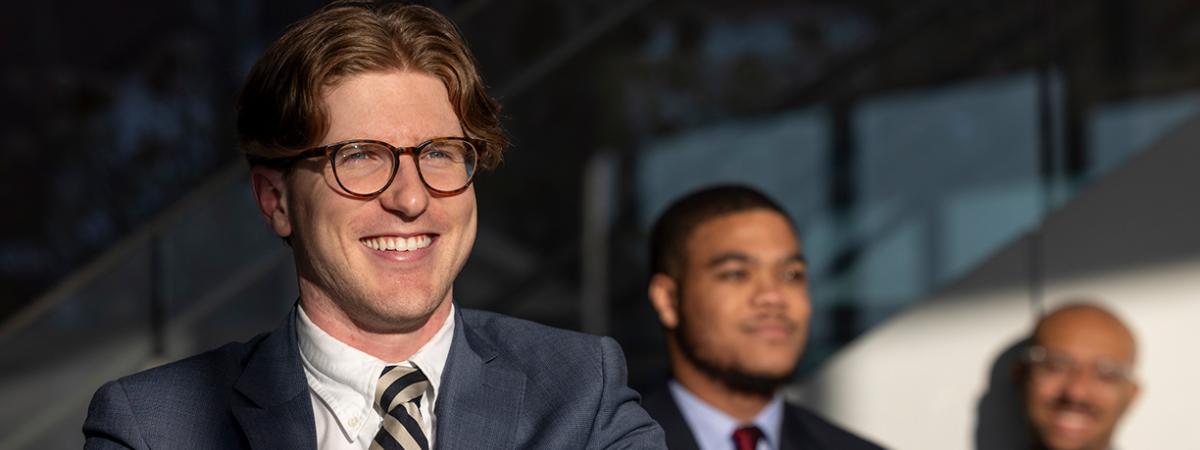 A smiling man with glasses and a striped tie in the foreground, with two colleagues in suits in the background.