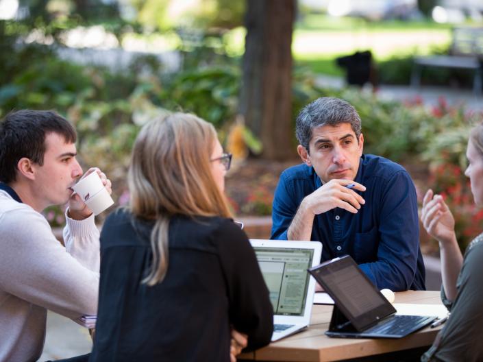 Professor Avi Cover sits at a table outside and talks with three individuals.