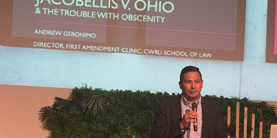 Director Andy Geronimo standing on a stage with a microphone and projector behind,presenting for a Continuing Legal Education seminar discussing the 1964 Jacobellis v. Ohio Supreme Court case in the Cleveland Heights Centrum Theater, where the events surrounding the case took place.