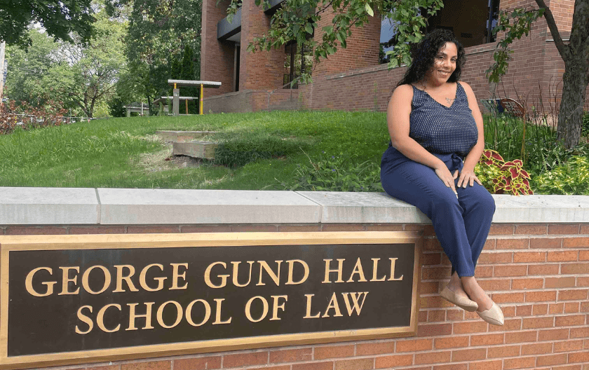 Heavenly Aguilar sitting on the wall in front of the law school with the sign reading "George Gund Hall School of Law" visible below her