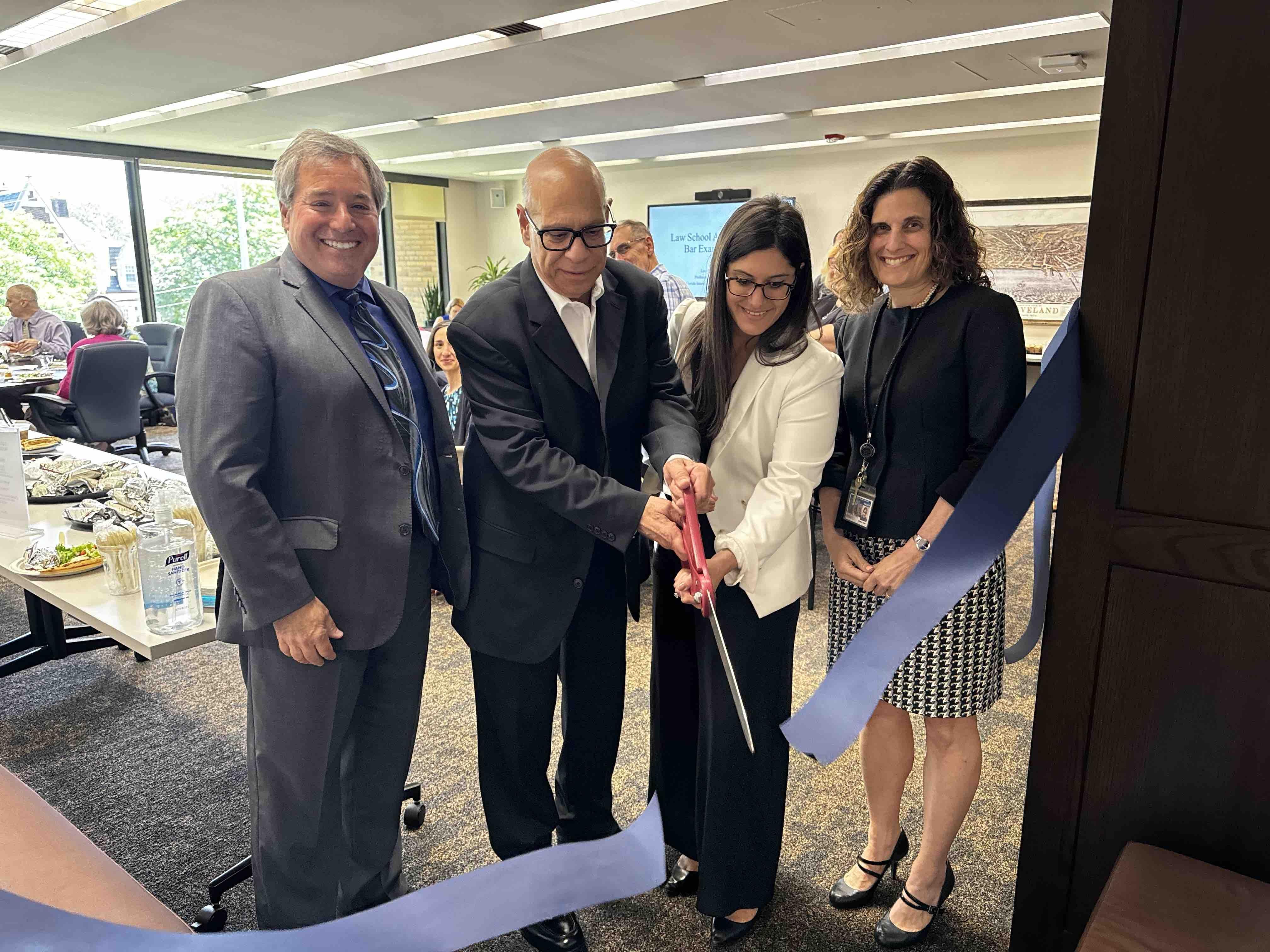 From left to right: Michael Scharf, George Simon, Stephanie Simon, Jessica Berg cutting a blue ribbon with giant scissors over the doorway of the newly renovated faculty lounge