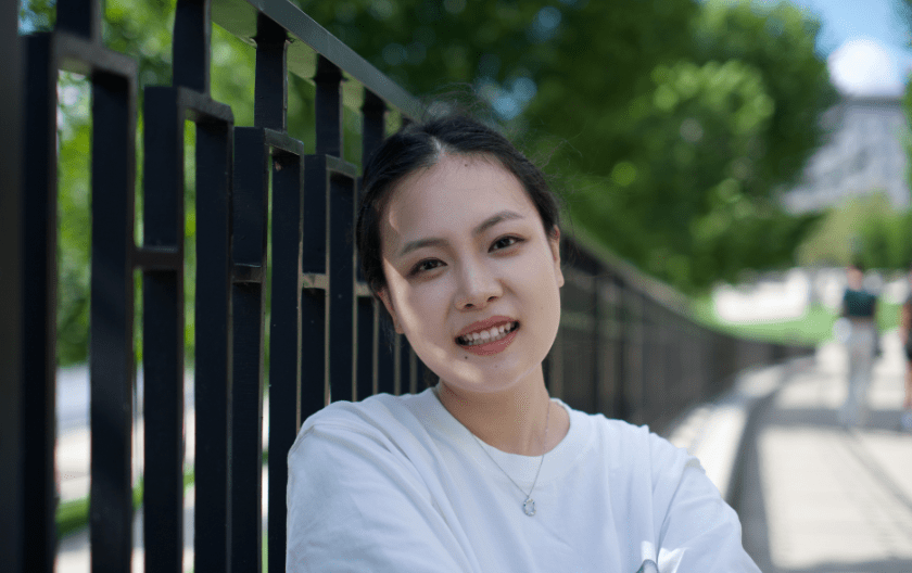 Xiaorui Han standing in front of a fence, smiling at the camera. There are trees behind the fence. 