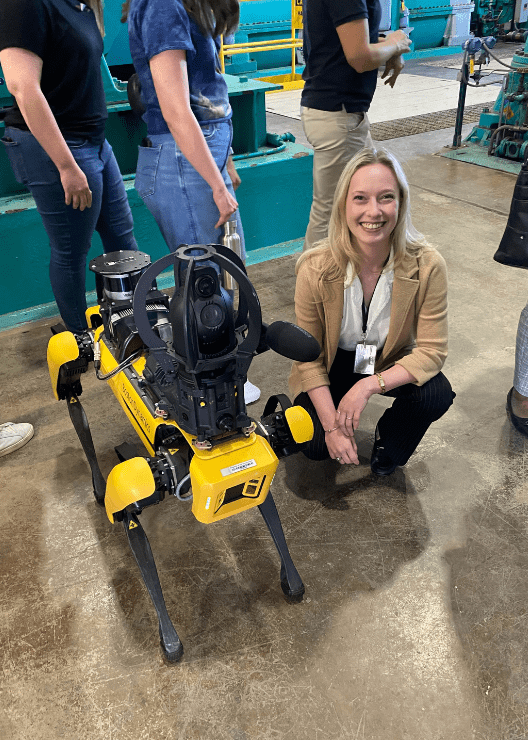 Annabelle Lincoln, smiling, crouched next to a robotic "dog" with other people standing and walking behind her.