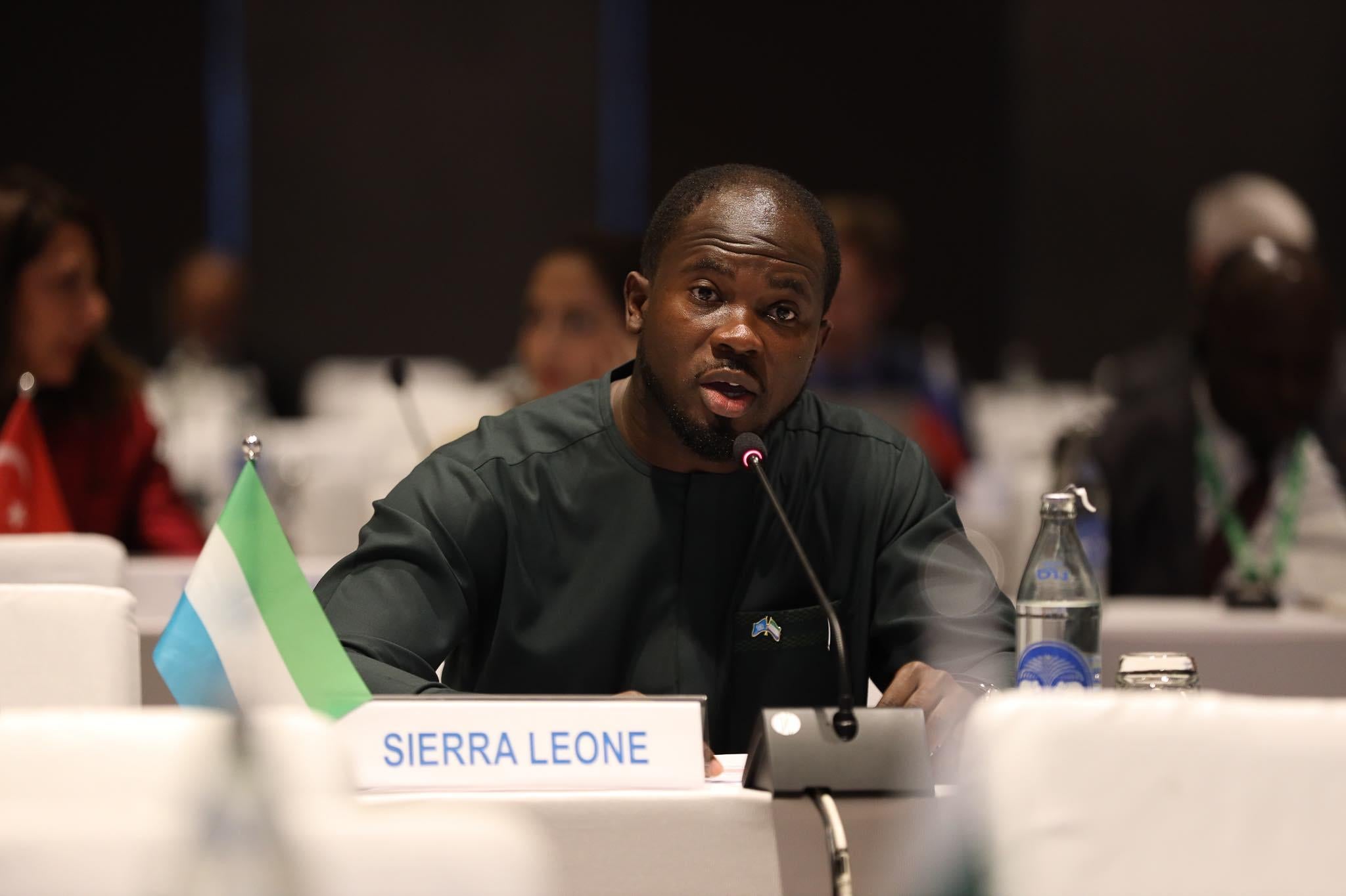 George Kamanda speaking into a microphone at a table with a plaque reading "Sierra Leone" in front of him