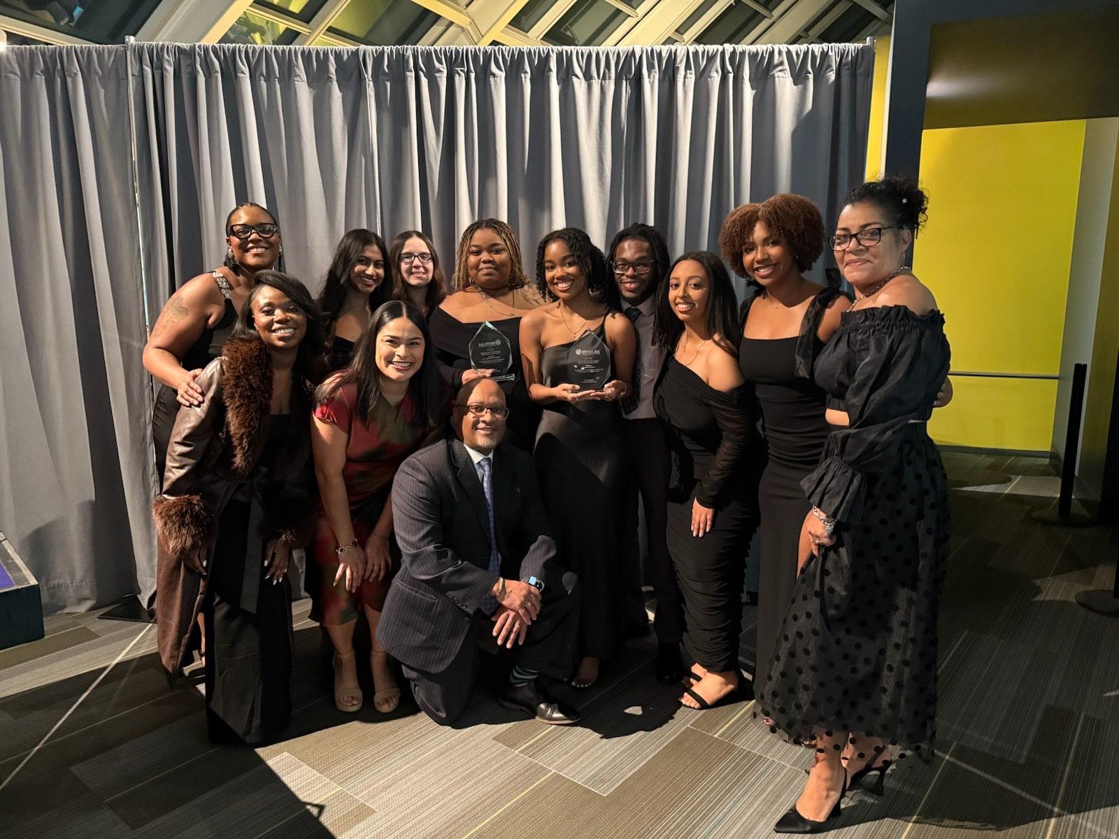 BLSA mock trial team and coaches, plus two additional students cheering on the team. They are posing for a photo while students hold trophies from the events. 