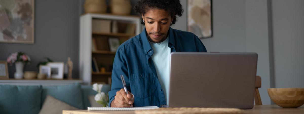 A young man sitting at a desk at home, writing in a notebook next to an open laptop.