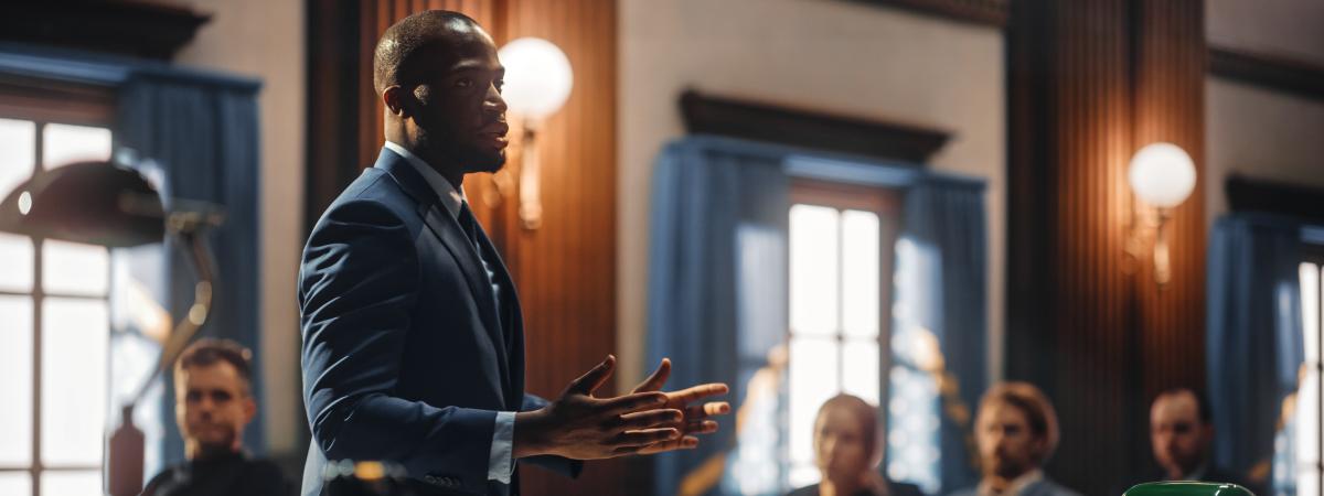 A male attorney in a formal suit standing and gesturing with open hands while addressing a room in a wood-paneled courtroom setting.