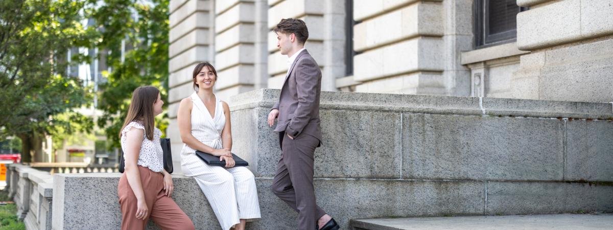 Three young professionals, two women and one man in a suit, conversing casually on the stone steps outside a large building.