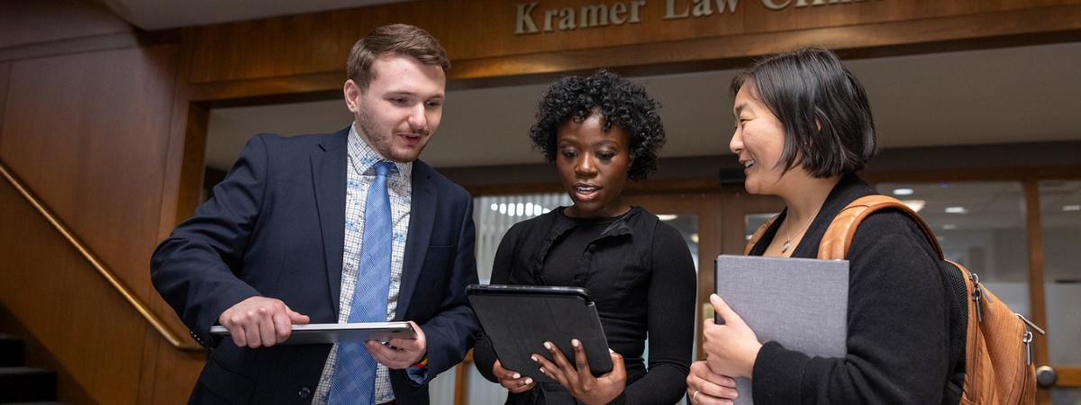 Three law students discussing a project while holding a tablet and laptop in a hallway.