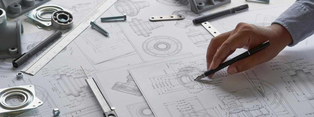 A person working on detailed engineering blueprints with drafting tools and mechanical parts scattered on the desk.