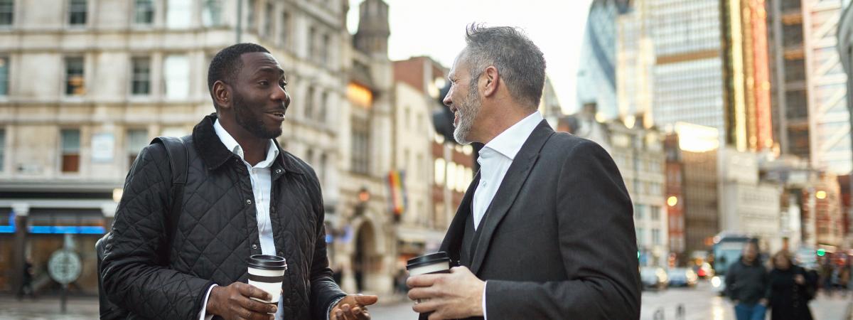 Two men chatting and holding coffee on a busy urban street with city buildings in the background.
