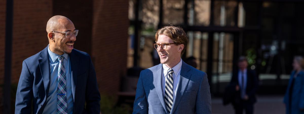 Two smiling men wearing business suits and glasses walking and talking together outdoors on a sunny day.