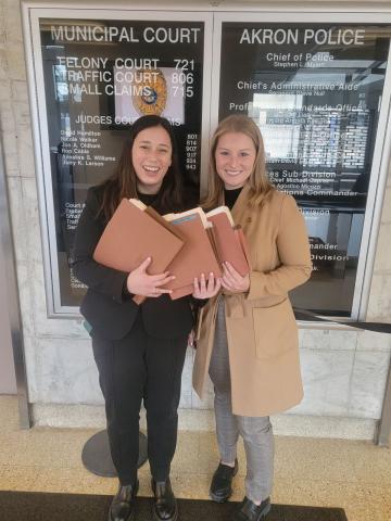 Two women with file folders posing in front of a sign for the Municipal Court and Akron Police Department.
