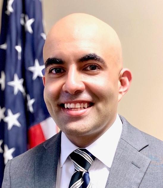 Sanjeev Bhasker headshot in front of an American flag. 