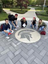 Students posing around a spartan helmet on the concrete walkway in the Quad