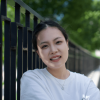 Xiaorui Han standing in front of a fence, smiling at the camera. There are trees behind the fence. 