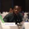 George Kamanda speaking into a microphone at a table with a plaque reading "Sierra Leone" in front of him