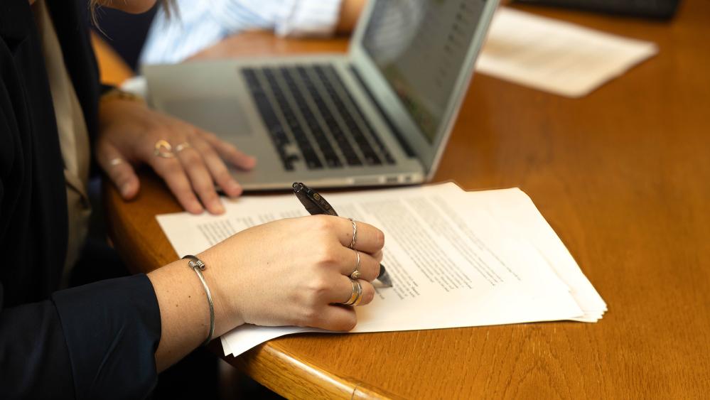 An individual uses a pen on a legal document with an open computer in the background.