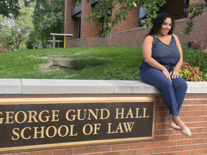 Heavenly Aguilar sitting on the wall in front of the law school with the sign reading "George Gund Hall School of Law" visible below her