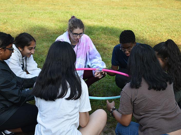 Seven students sitting outside on the ground in a circle holding a hoop
