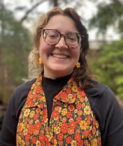 Headshot of Mailey Lorio. they have brown and blonde hair, glasses and are wearing a black turtle neck under an orange and yellow floral sleeveless top